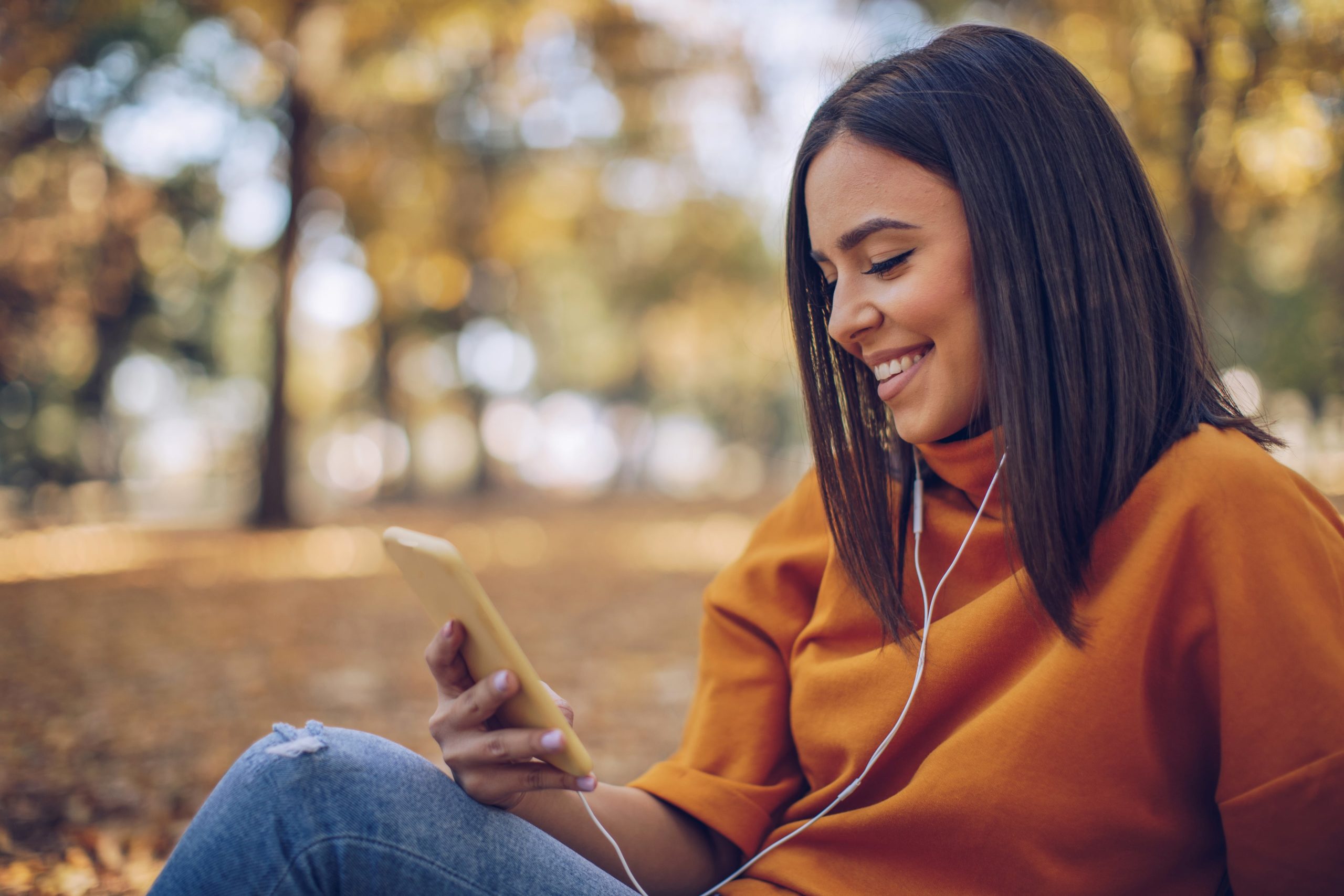 A woman in a park, smiling and looking at her phone, wearing earbuds, surrounded by autumn leaves. A moment of calm and connection.