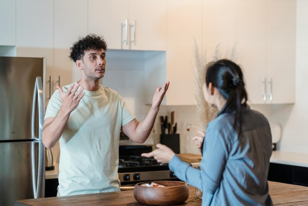 A distressed couple are across the kitchen from each other, having a painful conversation about infidelity and broken trust.