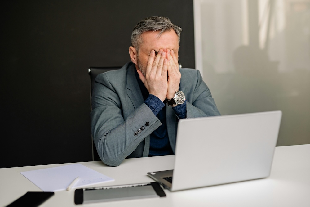 A man with his head in his hands at a laptop, depicting the brain fog and difficulty concentrating experienced during porn addiction withdrawal symptoms. This image represents the struggle with mental health during the early stages of recovery.