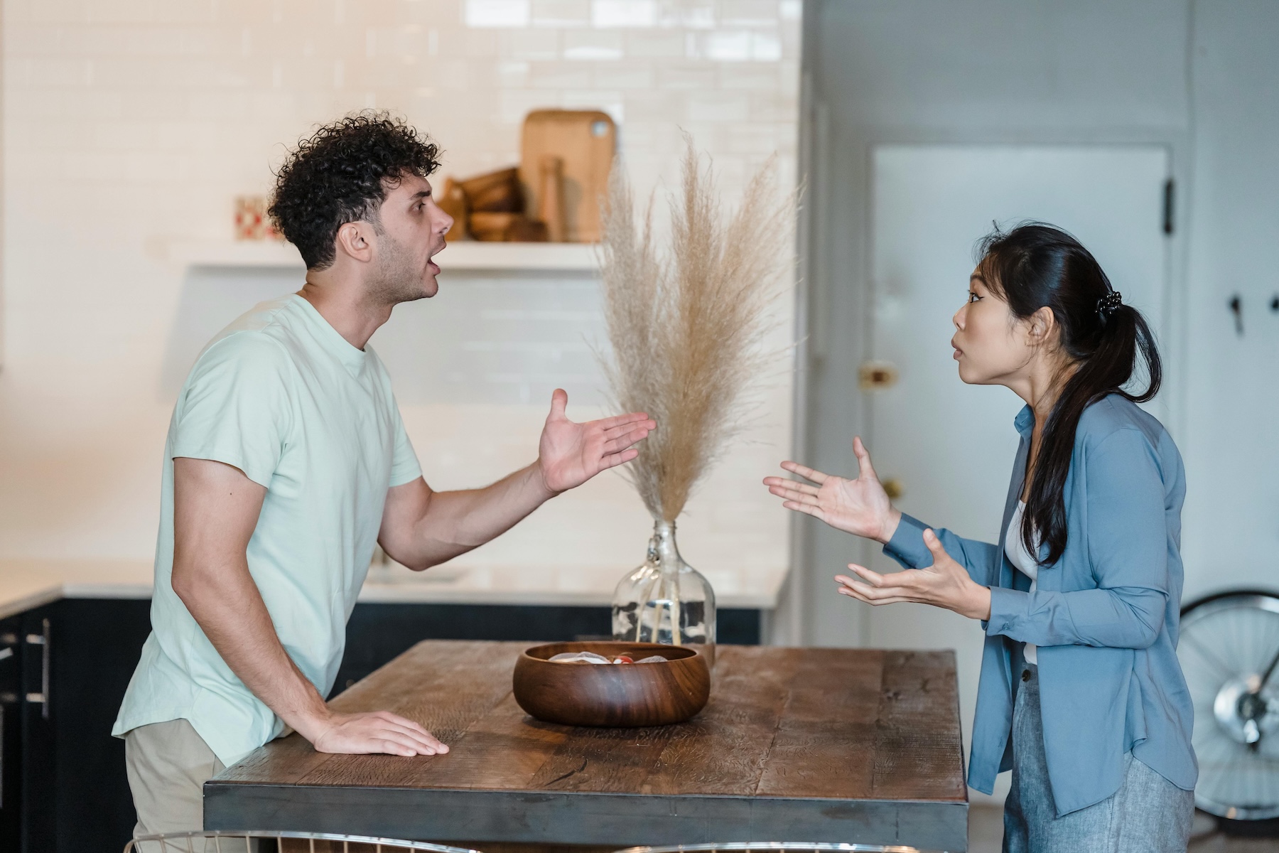 A distressed couple stands on opposite sides of their kitchen island, engaged in a heated argument, illustrating a sign of marriage conflict that may require couples therapy.