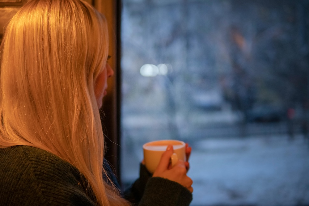 A woman peacefully looking out a window while holding a warm cup, representing a moment of nervous system regulation and resilience during recovery.