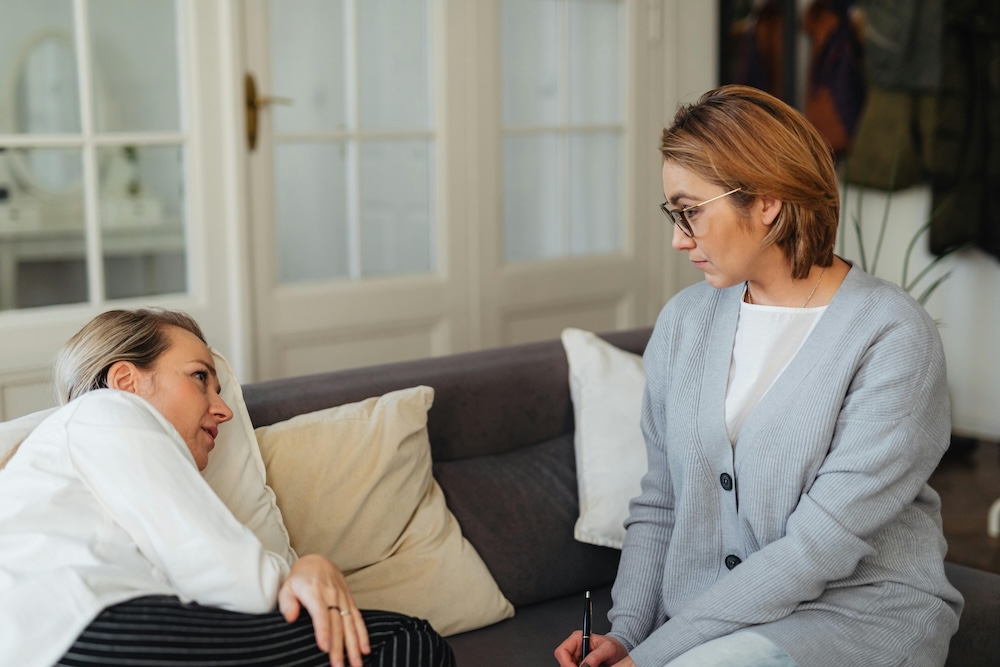 Two women engaged in a supportive and compassionate conversation on a sofa, illustrating the safety of a specialized trauma-informed counseling session.