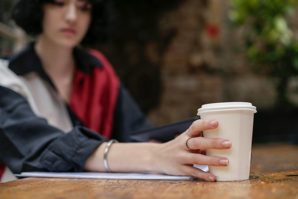 Hand with a ring reaches for a white takeaway coffee cup on a wooden table, with a blurred person in the background.