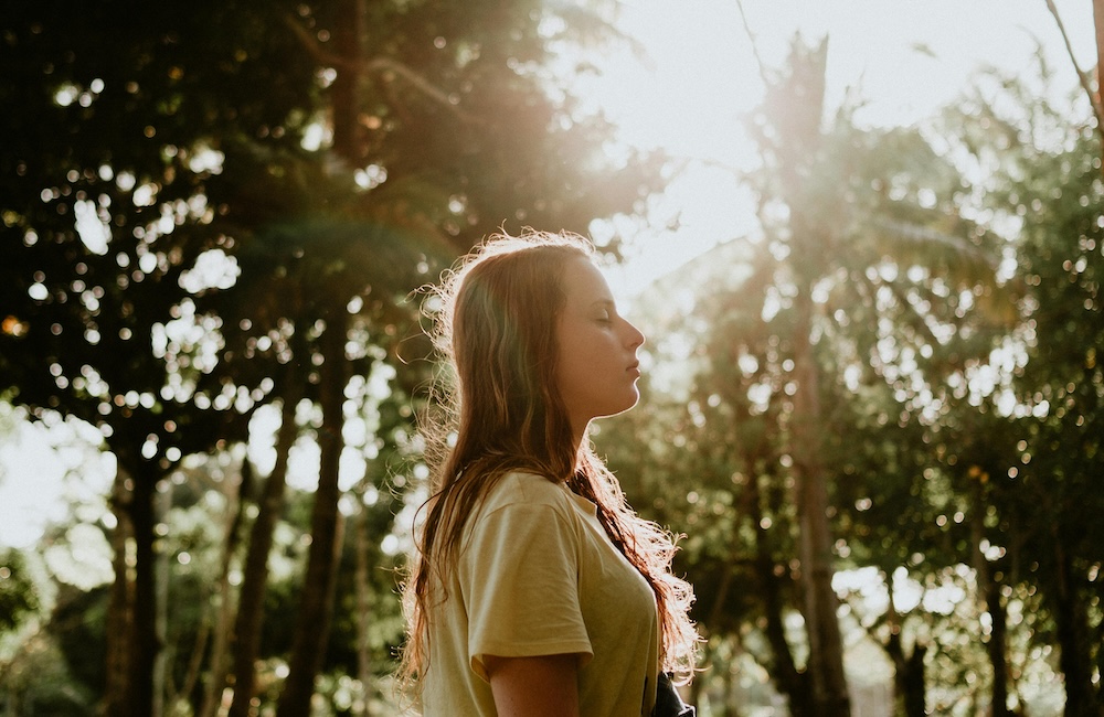 A woman standing in a peaceful outdoor setting with her eyes closed, symbolizing the shift from an activated nervous system to grounded inner knowing.