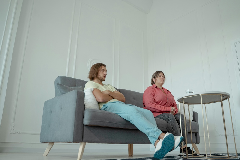 A couple sitting on a sofa with emotional distance, symbolizing the friction caused by an unequal mental load.