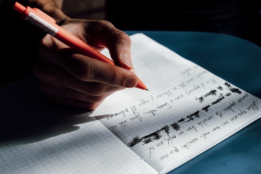 A person writing a letter by hand at a table — the work of naming what was chosen in restitution
