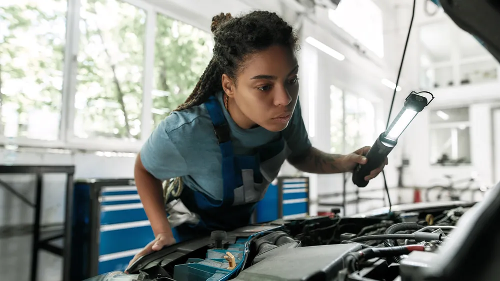 A mechanic shines a light under an open car hood, looking for what actually drives the engine