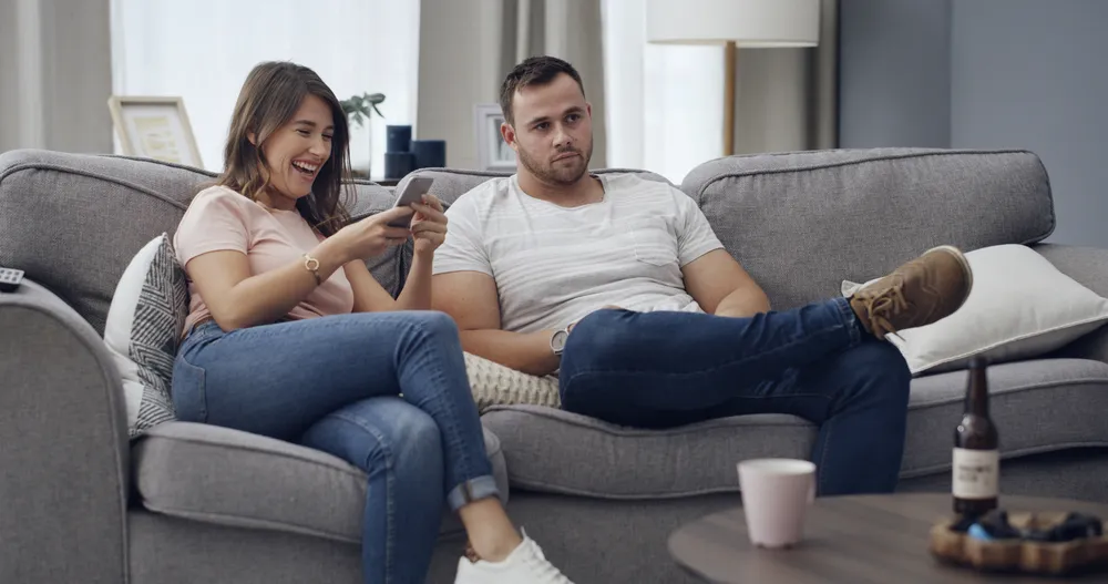 Couple on a sofa, one partner laughing at their phone while the other sits apart looking away