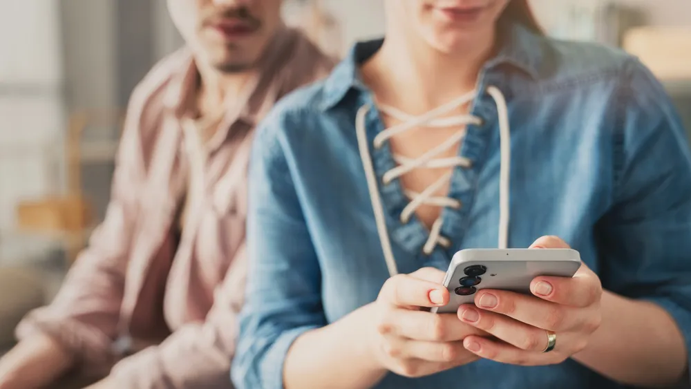 A person holds a phone with a wedding ring visible while their partner sits behind them.