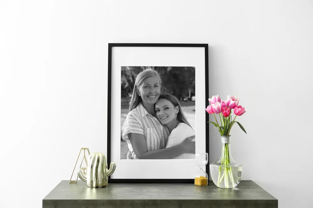 A framed black and white portrait of two women embracing, displayed on a shelf beside a vase of tulips.