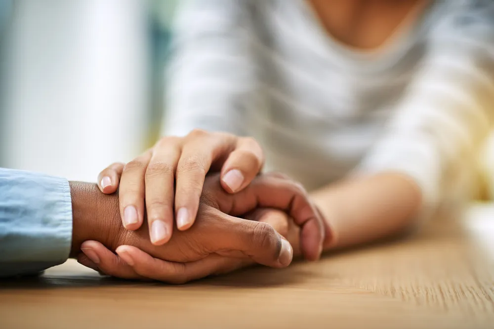 Close view of two people's hands held together on a wooden table, one hand resting over the other