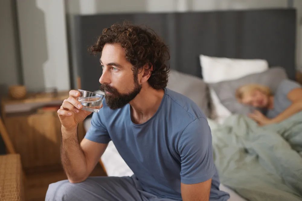 Person sitting on the edge of a bed at night holding a glass of water while their partner sleeps behind them