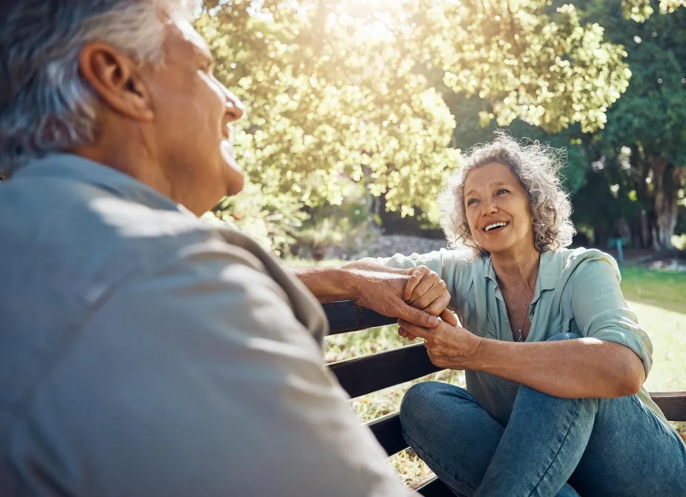 An older couple sitting on a park bench, turned toward each other and holding hands in conversation.