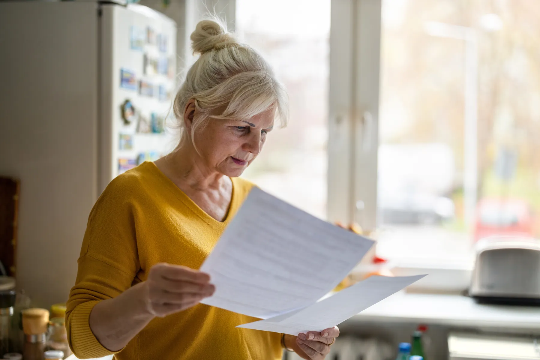 Woman standing in her kitchen reading a printed statement by the window, taking in what the numbers mean