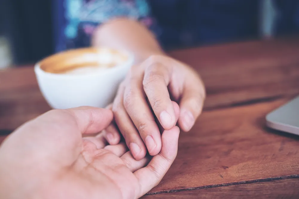 Two hands holding across a wooden table beside a coffee cup, one reaching toward the other rather than turning inward