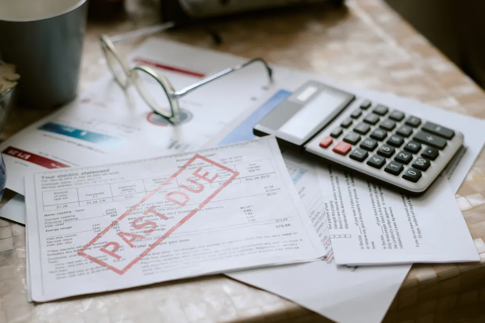 Past-due bills spread across a table with reading glasses and a calculator, the daily worry of whether there is enough