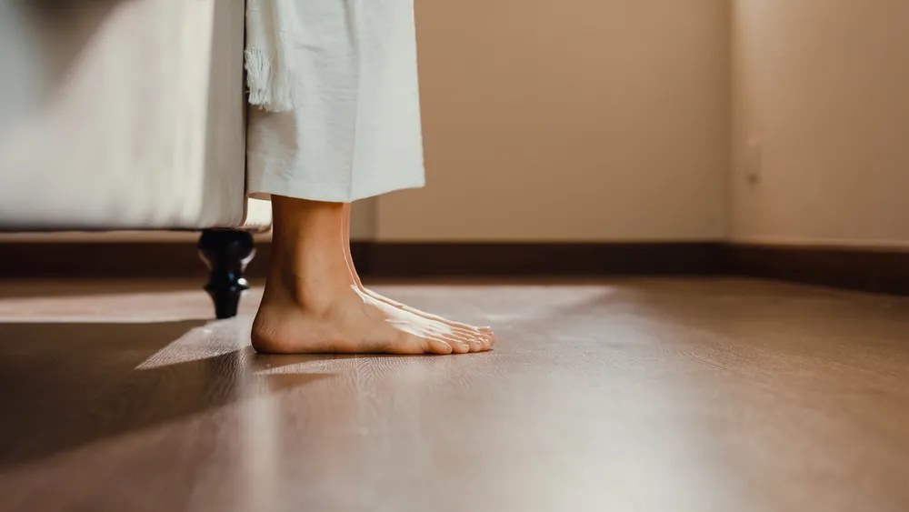 Bare feet planted flat on a hard floor beside a bed, a grounding step for regulating shame