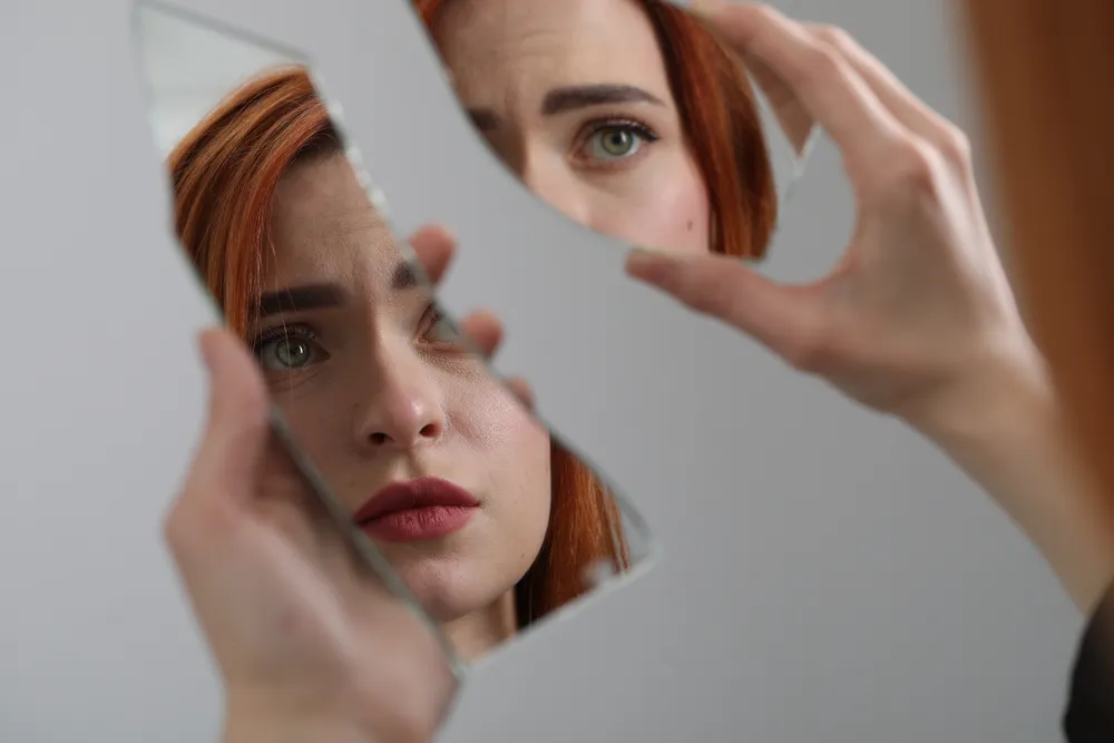 Broken mirror shards reflecting a person's face, an image of the shame spiral turning inward