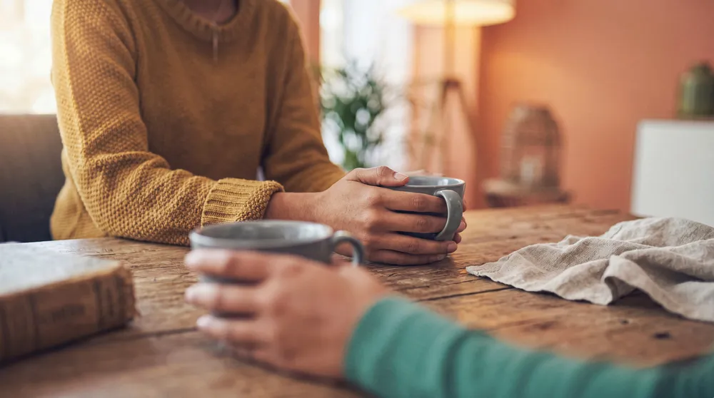 Two people holding coffee mugs across a wooden table in warm light, talking after telling a partner the truth
